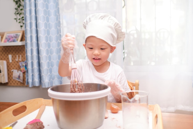 Lindo niño asiático de 4 años que se divierte preparando pasteles o panqueques disfruta el proceso mezcla la masa con un batidor en casa