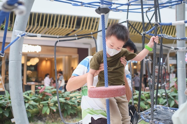 Lindo niño asiático con mascarilla lavable escalada en el gimnasio de la jungla en el patio interior pulbic con su padre durante el brote de covid-19, nuevo concepto de estilo de vida normal - Enfoque selectivo