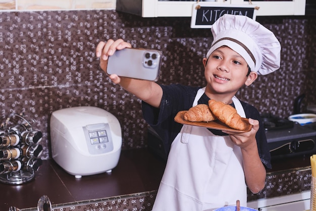 Foto lindo niño asiático con uniforme de chef tomándose selfie usando un smartphone para mostrar pasteles a las redes sociales