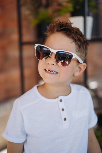 Lindo niño cauasiano en camiseta blanca y gafas de sol posando para la cámara y mostrando su diente perdido