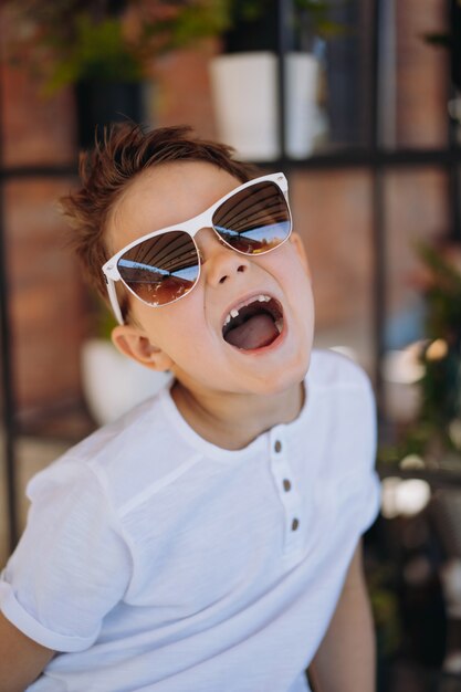 Lindo niño cauasiano en camiseta blanca y gafas de sol posando para la cámara y mostrando su diente perdido