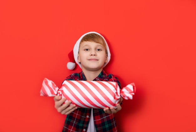 Lindo niño caucásico adolescente con sombrero de Santa sostiene una caja de regalo con forma de caramelo con sorpresa en el fondo rojo Copiar espacio para diseño o texto Plantilla de maqueta de Banner Navidad Año Nuevo