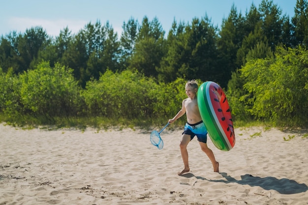 Lindo niño caucásico con anillo de goma y cazamariposas corriendo hacia el agua con risas Vacaciones en el lado del mar Infancia feliz Imagen con enfoque selectivo