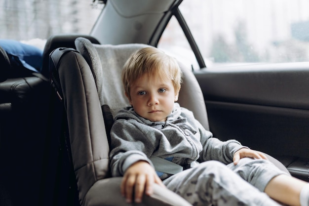 Lindo niño caucásico con cara triste sentado en un asiento de seguridad para niños en un coche