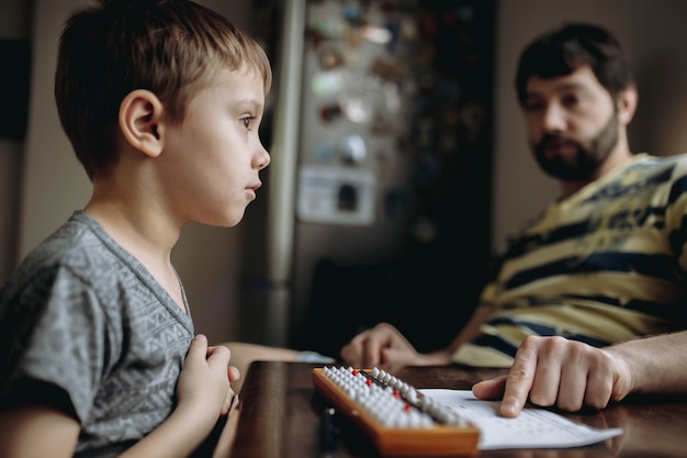 Lindo niño caucásico haciendo sus deberes de aritmética mental con el padre sentado junto a él en la mesa de la cocina. Foto de alta calidad