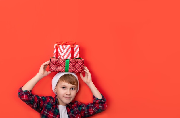 Lindo niño caucásico niño adolescente en cuadros cortos sostiene en la cabeza cajas de regalo sobre fondo rojo Copiar espacio para diseño o texto Banner Navidad Año Nuevo plantilla de maqueta