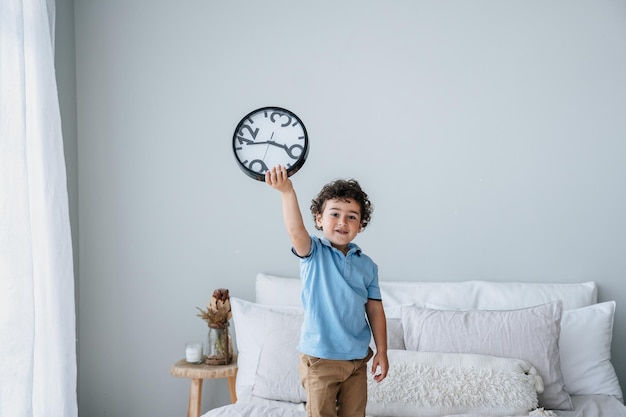 Lindo niño caucásico en polo azul con reloj de pared de pie en la cama temprano en la mañana levantándose Concepto de gestión del tiempo Niño español rizado mira a la cámara Niño pide pasar tiempo con los padres