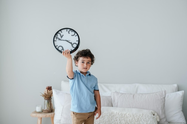 Foto lindo niño caucásico en polo azul sosteniendo el reloj de pared de pie en la mala madrugada levantándose concepto de gestión del tiempo niño español rizado mirando con confianza a la cámara con espacio vacío para el anuncio