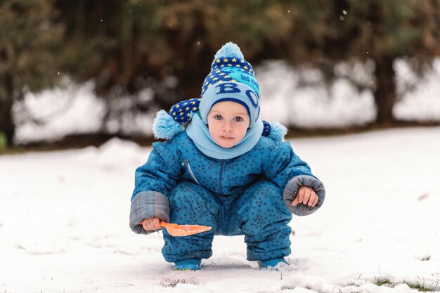 Lindo niño caucásico en ropa de abrigo y con bufanda y sombrero jugando en la nieve con pala pequeña.