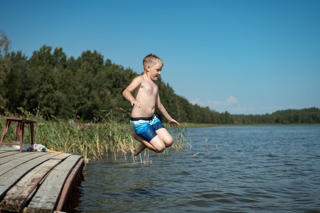 Lindo niño caucásico saltando desde el muelle de madera sumergiéndose en el lago en el campo