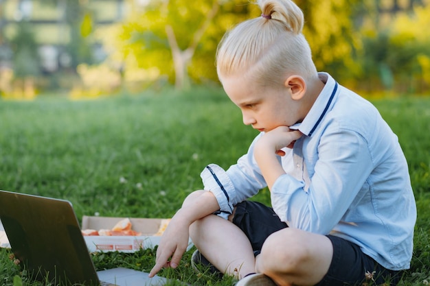 Lindo niño caucásico sentado en el parque de hierba con computadora portátil