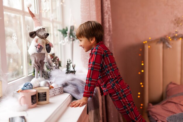 Lindo niño curioso en pijama a cuadros, mirando a través de una ventana, esperando con impaciencia a Santa y regalos