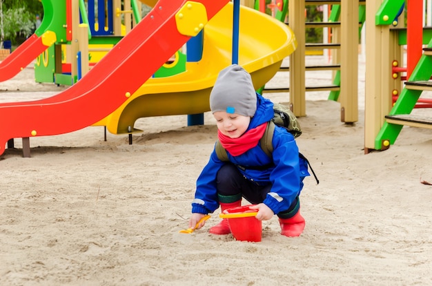 Foto lindo niño de dos años en una chaqueta azul y una bufanda roja juega en un patio brillante con una cuchara y un balde