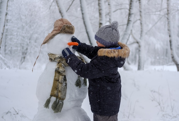 Lindo niño europeo y el gracioso muñeco de nieve en el bosque nevado en un día de invierno en la nieve.