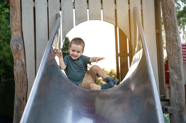 Un lindo niño feliz de dos o tres años bajando por el tobogán en el patio de recreo en un día de verano Niño activo jugando afuera Enfoque selectivo