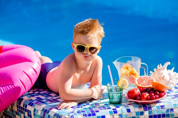 lindo niño con gafas de sol y traje de baño bebe limonada junto a la piscina.