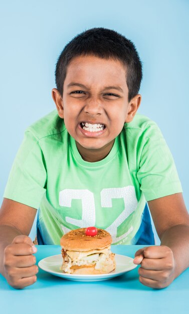 Lindo niño indio comiendo hamburguesa, pequeño niño asiático y hamburguesa, sobre fondo azul.