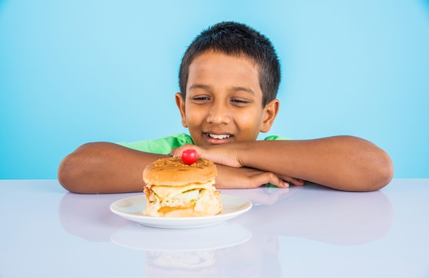 Lindo niño indio comiendo hamburguesa, pequeño niño asiático y hamburguesa, sobre fondo azul.