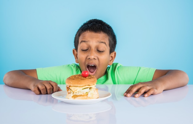 Lindo niño indio comiendo hamburguesa, pequeño niño asiático y hamburguesa, sobre fondo azul.