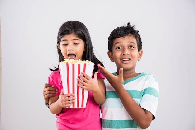 Lindo niño indio feliz y niña o hermanos comiendo palomitas de maíz y viendo la televisión mientras está sentado en una silla