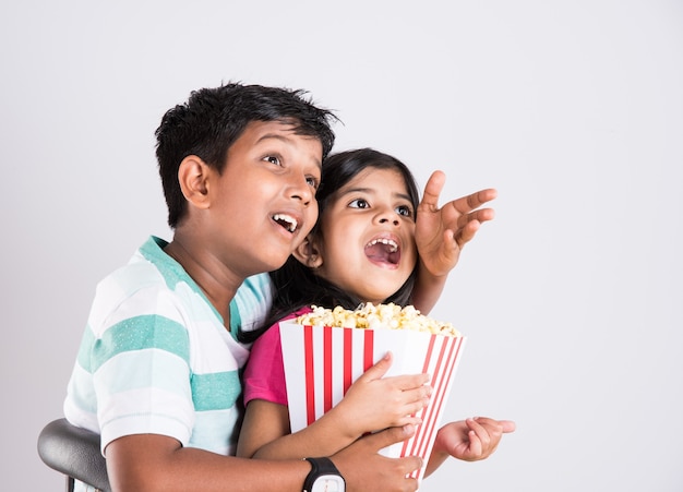 Lindo niño indio feliz y niña o hermanos comiendo palomitas de maíz y viendo la televisión mientras está sentado en una silla