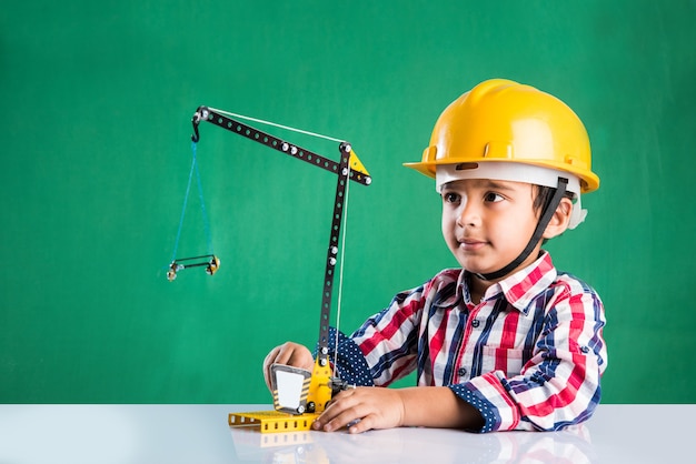 Lindo niño indio jugando con grúa de juguete con sombrero de construcción amarillo o casco, concepto de infancia y educación, aislado sobre pizarra verde