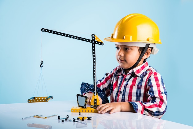 Lindo niño indio jugando con grúa de juguete con sombrero de construcción amarillo o casco, concepto de infancia y educación, aislado sobre pizarra verde