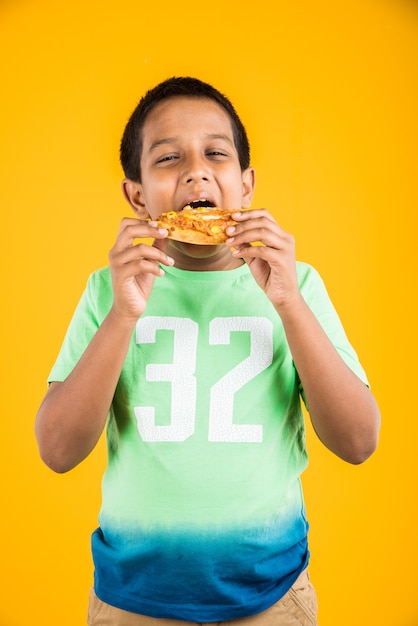 Lindo niño indio o asiático comiendo sabrosas hamburguesas, sándwiches o pizzas en un plato o caja. Permanente aislado sobre fondo azul o amarillo.