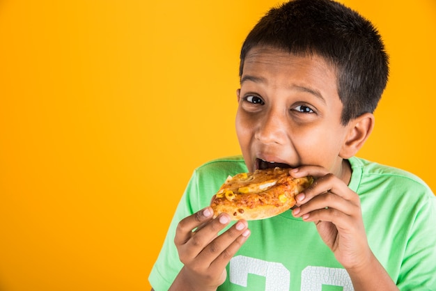 Lindo niño indio o asiático comiendo sabrosas hamburguesas, sándwiches o pizzas en un plato o caja. Permanente aislado sobre fondo azul o amarillo.