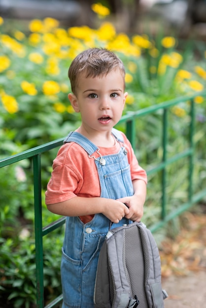 un lindo niño mira a la cámara sostiene una cartera en sus manos vestido con un mono de mezclilla