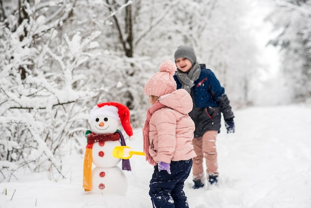 Lindo niño y niña construyendo muñeco de nieve en bosque blanco de invierno