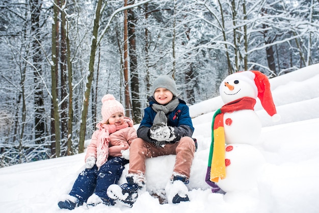 Lindo niño y niña construyendo muñeco de nieve en bosque blanco de invierno