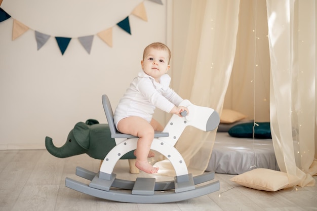 Lindo niño o niña en una luminosa habitación infantil con un wigwam y un elefante en un traje de algodón blanco balanceándose en un caballito de madera