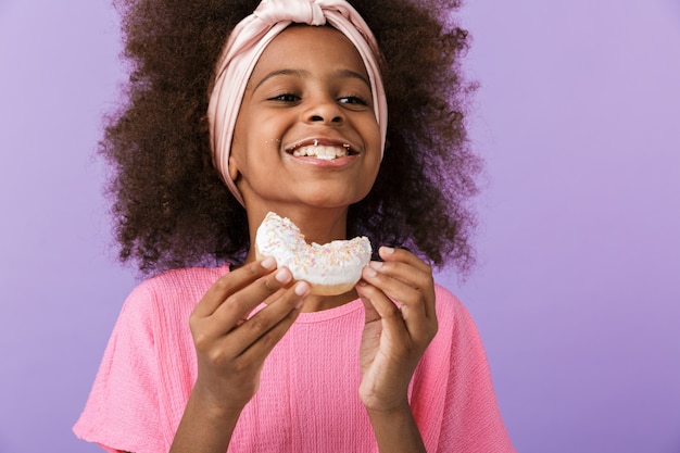 Lindo niño optimista joven africana posando aislado sobre pared púrpura, come donut.