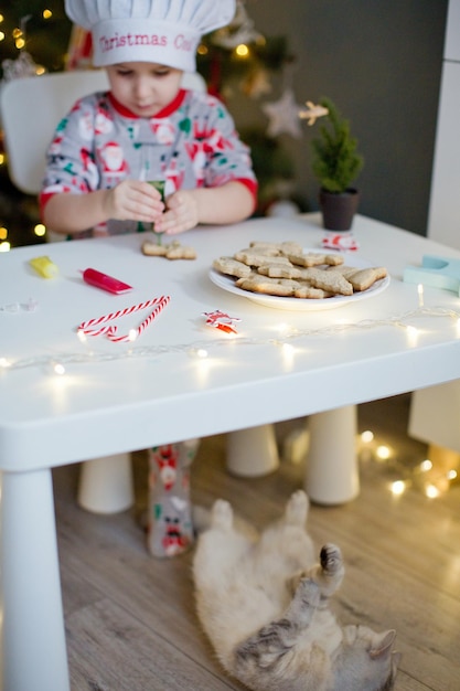 Lindo niño pequeño decora galletas de Navidad con glaseado de colores en una mesa blanca cerca del árbol de Navidad con luces Gato gris y niño en un fondo de Navidad Concepto de cocina de Navidad