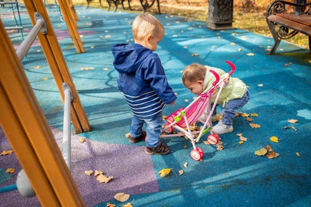 lindo niño pequeño y hermosa niña de un año juegan en un cochecito de juguete para una muñeca en el parque de otoño