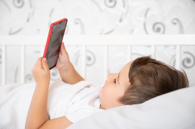 Lindo niño pequeño mirando smartphone en la cama. Concepto de dependencia de Internet.