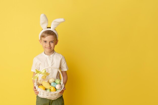 Lindo niño pequeño con orejas de conejo sosteniendo canasta de Pascua sobre fondo amarillo