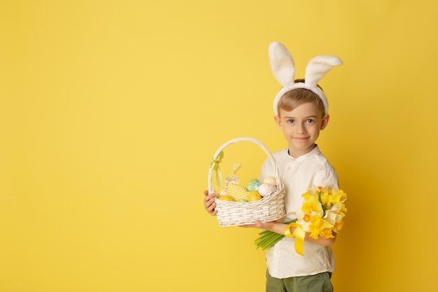 Lindo niño pequeño con orejas de conejo sosteniendo canasta de Pascua sobre fondo amarillo