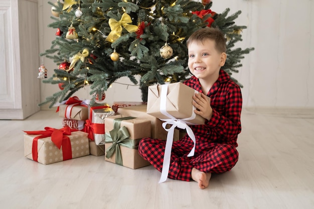 lindo niño pequeño en pijama rojo con cajas de regalo sentado bajo el árbol de navidad en casa