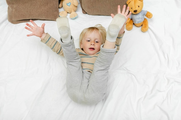 Lindo niño pequeño rubio caucásico niño niño pequeño jugando divirtiéndose acostado en la cama levanta las piernas en casa en la mañana soleada se despierta en su habitación momentos felices de la infancia