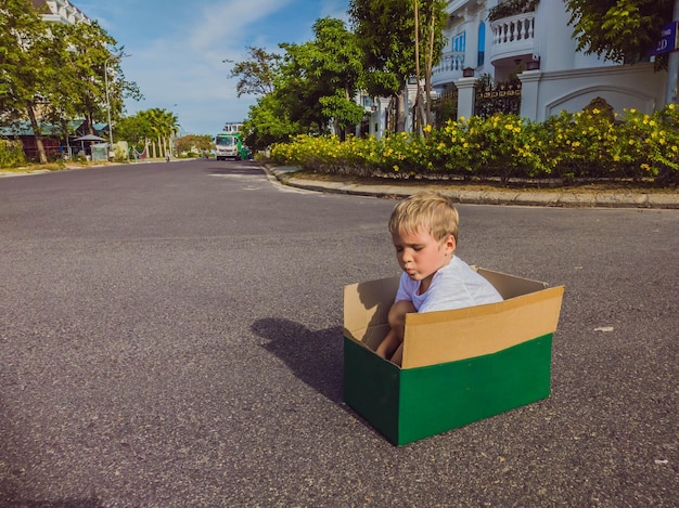 Lindo niño pequeño sentado dentro de una caja de cartón en una calle silenciosa en un moderno y cómodo pueblo rural Infancia y concepto de mudanza