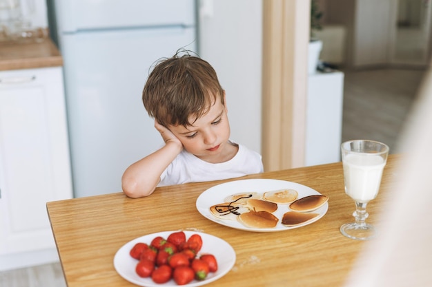 Lindo niño pequeño sentado con puncakes con bayas en la cocina de la casa