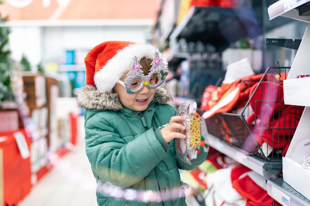 Lindo niño preescolar con sombrero de Navidad y máscara eligiendo decoración de año nuevo en el supermercado. Familia comprando regalos de Navidad.