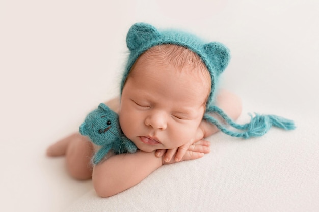 Un lindo niño recién nacido en los primeros días de vida duerme desnudo contra un fondo de tela blanca gorra azul azul con orejas como un oso oso de juguete en las manos foto de alta calidad