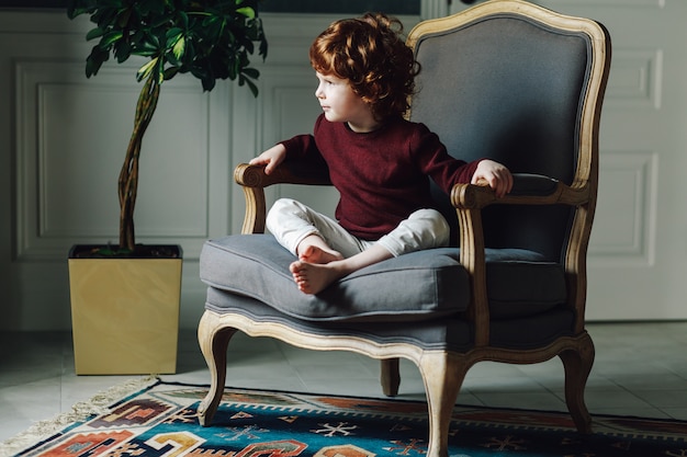 Lindo niño rizado sentado relajado en un cómodo sillón y mirando a otro lado