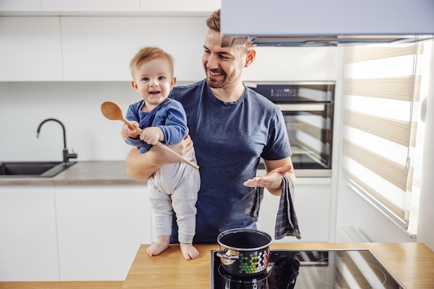 Lindo niño rubio de pie sobre la encimera de la cocina y sosteniendo una cuchara mezcladora. Su padre sosteniéndolo.