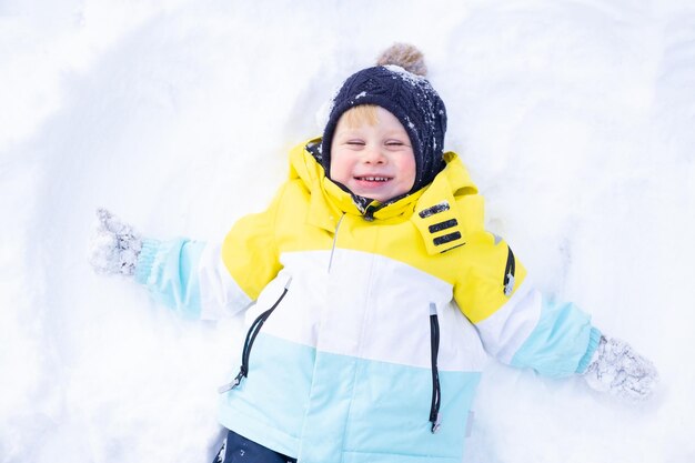 Lindo niño sonriente acostado en un ventisquero, divirtiéndose, riendo al aire libre en el bosque de invierno