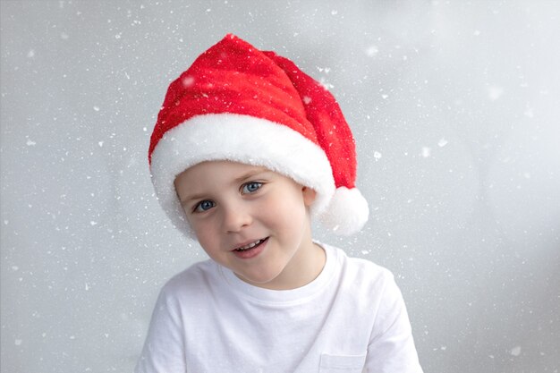 Lindo niño sonriente en una gorra roja de Navidad está feliz de fondo claro con nieve Concepto de Navidad