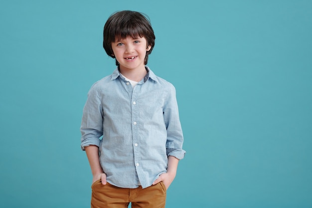 Lindo niño sonriente manteniendo las manos en los bolsillos de los pantalones marrones